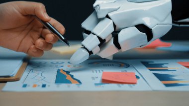 Bir insan eli ve robot el iş haritaları üzerinde birlikte çalışır, veri eğilimlerini analiz eder ve iş yerindeki teknoloji ile insan yaratıcılığının kaynaşmasını gösterir. Noogenesis