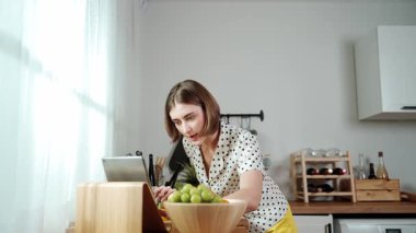 Kafkasyalı anne, modern mutfakta dizüstü bilgisayarda yemek tarifine bakarken salata yapmak için yemek hazırlıyor. Yetenekli ev bekçisi taze sebze kullanarak kahvaltı hazırlıyor. Pedagoji.