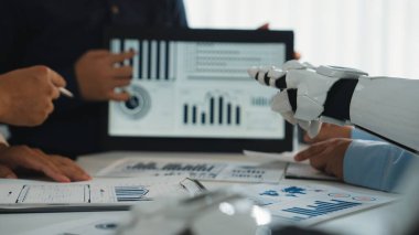 Modern bir iş toplantısı sahnesi, insan eliyle ekip çalışması ve dijital tablette dinamik grafikleri ve grafikleri analiz eden bir robot kol yenilikleri vurguluyor.
