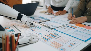 Modern bir çalışma alanı sahnesinde robot ve insan birlikte çalışıyor, çeşitli veri tabloları ve raporlarını analiz ediyor, iş dünyasındaki yenilik ve teknolojiyi somutlaştırıyor. Noogenesis