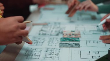 Renkli kalemlerle dolu renkli bir beyin fırtınası oturumunda masa üzerinde detaylı mimari planlar yapan çeşitli profesyonellerin ellerinin yakın çekimi. SACTR
