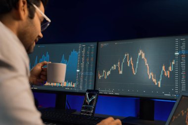 Zeki tüccar işadamı kahve fincanı içiyor, neon ışıklı ofisteki arama pazarında yüksek karlı finans teknolojisiyle bilgisayar üzerinde iki dinamik borsa yatırım ekranını analiz ediyor. Tahmin et..