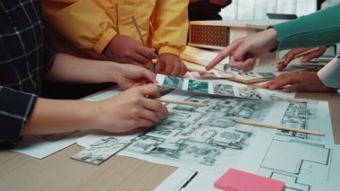 Bir grup birey bir masada mimari planları tartışıyor ve analiz ediyor. Sahne, kapalı alanda yaratıcı bir tasarım sürecine aktif olarak dahil olan elleri vurguluyor. SACTR