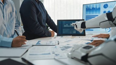 Modern bir ofis kurulumu, insan katılımcıların ekranda gösterilen veri ve mali raporları analiz ederek robotik bir kolla işbirliği yaptıkları bir iş toplantısı gösteriyor.