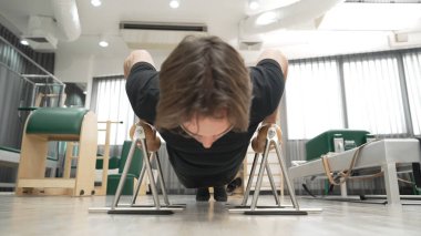 Müşteri, bir spor salonunda üst vücut gücünü, dengeyi ve çekirdek bağını geliştirmek için pilates egzersizi sırasında tutamaklarını kullanarak şınav çekiyor. Adam egzersiz yapıyor ve şınav çekiyor. Alışkanlık.