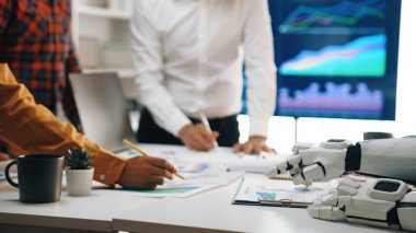 Çeşitli bir ekip, verileri analiz etmek, stratejileri tartışmak ve gelecek gelişimi için plan yapmak amacıyla modern ofis ortamında, teknoloji ve robot yardımından yararlanmak için işbirliği yapmaktadır.