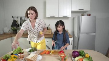 Zeki beyaz anne ve Asyalı kız birlikte yemek pişiriyor ve sebze doğruyor ya da akşam yemeği için salata hazırlıyor. Mutlu anne ve kız taze yemekle sağlıklı yemek yapıyorlar. Eğitim konsepti. Pedagoji.