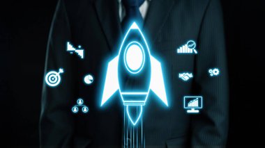 Takım elbiseli bir işadamı, büyümeyi ve yeniliği sembolize eden, girişimciliğin ve başarının özünü vurgulayan, parlayan bir dijital roket ikonu ile güvenle ayakta duruyor. Dörtgen