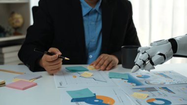 Bir iş adamı, veri tabloları ve grafiklerini analiz ederken robotik bir kol ile işbirliği yapar, modern çalışma ortamlarında teknolojinin entegrasyonunu vurgular. Noogenesis