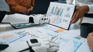 İş toplantısına katılan bir robot ve insan, konferans masasında çizelgeler ve veri analizleri sergiliyor, iş yerinde gelişmiş işbirliği ve teknoloji gösteriyor. Noogenesis