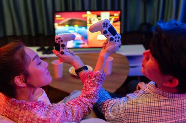 joystickli elleri çapraz oynayan bir çift arka tarafta TV ekranında dövüş oyunu oynuyor, modern rahat evinde neon ışıklı oturma odasında kanepesinde oturuyor. İnfobahn.