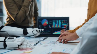 Renkli çizelgeler ve dijital aletler kullanarak veri içgörülerini analiz etmek için toplantının ortasında insan eli ve robot kolların işbirliği yaptığını gösteren modern bir çalışma alanı. Noogenesis