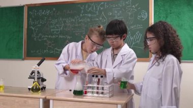 Amerikalı kız deney kabını solüsyonla tutuyor ve deney için hazırlanıyor. Meraklı mutlu ilkokul öğrencisi laboratuarda sıvı döküp karatahtaya kimyasal teori ile karıştırıyor. Pedagoji.