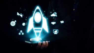 Parlayan bir roket ikonunu dijital alanda yenilik ve büyümeyi temsil eden çeşitli iş simgeleriyle birlikte gösteren akıllı telefonu tutan bir elin yaratıcı görüntüsü. Dörtgen