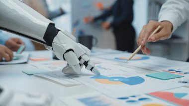 Bir masadaki renkli çizelgeleri ve grafikleri analiz eden robotik bir elin gelecekteki bir sahnesi teknoloji ve insan çabası arasındaki işbirliğini temsil ediyor. Noogenesis