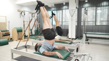 Müşteri, spor salonundaki yenilikçi bir makinede kayışla ters bacak kaldırma hareketi yapıyor. Çekirdek bağlantısını, dengesini ve esnekliğini geliştirmek için ters takla atan iki mutlu antrenör. Alışkanlık.