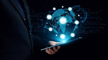 Bir işadamı, modern bir iş ortamında küresel bağlantı, bilgi paylaşımı ve teknolojik yeniliği temsil eden gelecekçi bir dijital arayüzü gösteren bir tablet tutuyor. Dörtgen