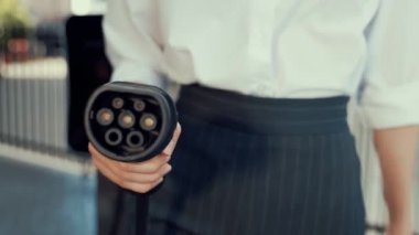 İlerici iş kadını şehir merkezinde dolaşmadan önce şarj cihazından elektrikli arabasına şarj prizi takıyor. Çevre dostu şarj edilebilir araba, sürdürülebilir ve temiz enerjiyle çalışıyor..