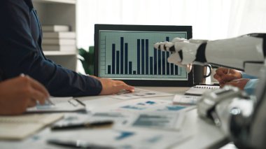 Ekip çalışmalarını ve yenilikleri vurgulayarak ekranda sunulan verileri insan eli ve robotik kolun analiz ettiği ortak bir iş toplantısını gösteren dinamik bir sahne. Noogenesis