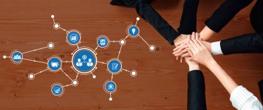 Çeşitli bir iş ekibi ahşap bir masada birlik ve işbirliği gösterisinde el ele tutuşuyor. Çevrelerindeki dijital simgeler, iş yerlerindeki stratejiyi, iletişimi ve yeniliği sembolize ediyor. Dostluk