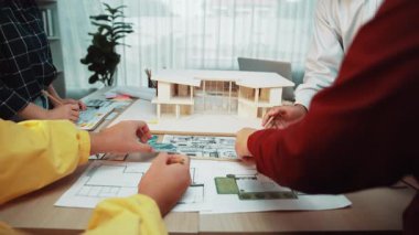 Çeşitli bir ekip, yenilikçi fikirler üzerinde çalışırken modern ofis ortamında mimari tasarımlar, planlar ve fiziksel bir model üzerinde işbirliği yapıyor. SACTR