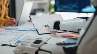 Bir robot el, modern bir ofis masasında çeşitli çizelge ve raporlarla etkileşime geçerek veri analizlerinde teknoloji ve takım çalışmasının harmanlanmasını gösterir. Noogenesis