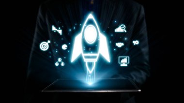 Takım elbiseli bir işadamı, yenilik, büyüme ve iş başarısını temsil eden dijital teknoloji simgeleriyle çevrili parlayan bir roket grafiğini gösteren bir tablet tutuyor. Dörtgen