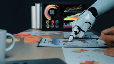 Bir insan eli, modern bir ofiste veri tabloları ve raporlarını incelerken robotik bir elle işbirliği yapar. Görüntü yenilik ve teknoloji entegrasyonunu gösteriyor. Noogenesis