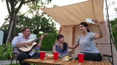 Baba gitar çalarken anne ve kızı bahçede kamp yapıyor. Ebeveynler, genç nesil çapraz kuşak farklılıklarıyla zaman geçirmek için açık hava kamp aktivitelerini kullanıyorlar. Uyumsuzluk.