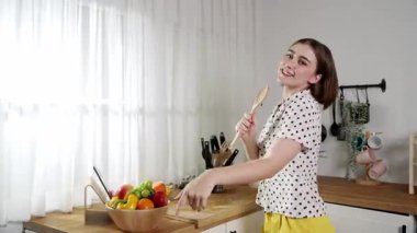 Enerjik beyaz anne kahvaltı için salata pişirirken ve yemek pişirirken elinde kepçeyle şarkı söylüyor. Sağlıklı yemek yapmaya hazırlanırken müziğe yönelen mutlu ev sahibi. Pedagoji.