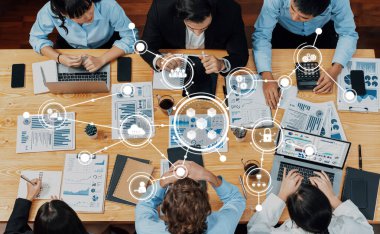 Çeşitli bir iş ekibi, iletişim ve analizi temsil eden dijital simgelerle çevrili ve üretken strateji planlamasını geliştiren ahşap bir masada işbirlikçi bir tartışmaya girişiyor. Dostluk