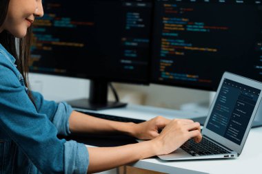 IT geliştiricisinde gülümseyen genç Asyalı dizüstü bilgisayarında kodlama programı verileriyle kotlu tişört giyerek çevrimiçi bilgi yazıyor. Güvenlik analizi iki ekran ile çevrili. Taktik.