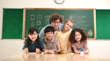 Kafkas profesyonel öğretmen sınıfta çeşitli sevimli öğrencilere sarılırken ve kameraya bakarken oturuyor. Sınıfta dikilirken kameraya gülümseyen bir grup çok kültürlü çocuk. Pedagoji.