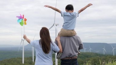 İlerici mutlu aile kavramı rüzgâr türbini çiftliğinde eğleniyor. Rüzgar türbini jeneratörüyle gelen elektrik jeneratörü kırsal bölgede tepe ve ufukta dağ var..