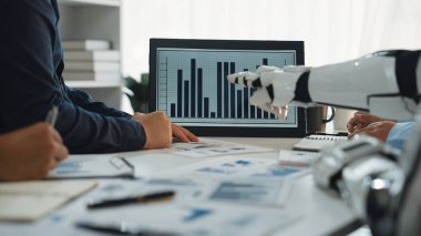 Robot bir elin ekranda veri analiz tablosuyla etkileşime geçtiği, iş yerindeki teknoloji ve işbirliğini vurgulayan modern bir iş toplantısı. Noogenesis