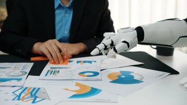 Bir profesyonel, bir robotik kol ile işbirliği yapar, masadaki finansal grafikleri ve çizelgeleri inceler, modern iş analizlerinde teknolojinin entegrasyonunu gösterir.