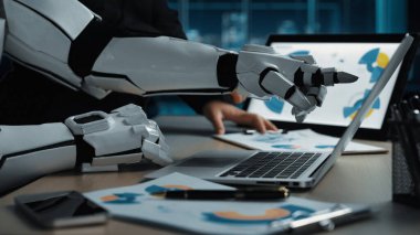 Yenilikçi çalışma alanlarında teknoloji ve takım çalışmasının yakınlaşmasını sembolize eden, dizüstü bilgisayarla uğraşan bir robotun modern ofis sahnesi. Noogenesis