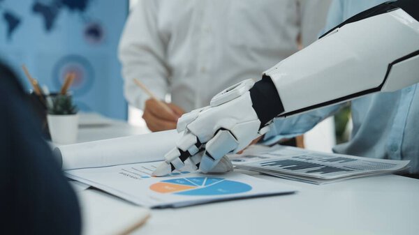 A robotic hand interacts with business charts and graphs during a team meeting in a modern office setting, showcasing the rise of technology in workplace collaboration.Noogenesis