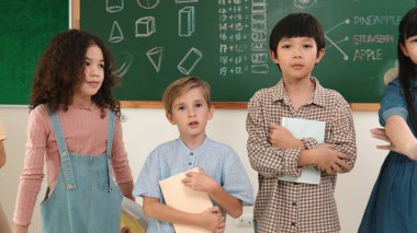 Çok kültürlü bir ilköğretim öğrencisi, günlük giysiler içinde sınıfta el sallarken kameraya bakıyor. Çeşitli çocuklardan oluşan bir grup not defteri tutuyor ve sınıftaki tahtaya gülümsüyor. Pedagoji.