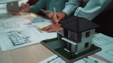 Çeşitli bir ekip mimari planlar üzerinde işbirliği yapar ve proje stratejilerini çalışma masasının üzerine dizilmiş planlar ve model bir evle tartışır. SACTR