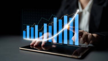 Profesyonel bir el tablet ekranında dijital bir grafikle etkileşime geçerek, modern ofis ortamındaki finansal büyüme eğilimlerini göstererek veri içgörülerini vurguluyor. Etki