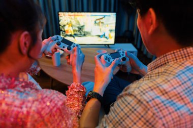 Kontrollü joystickli birkaç oyuncu arka tarafta televizyon ekranında dövüş oyunu oynuyor, modern rahat evinde neon ışıklı renkli oturma odasında koltukta oturuyor. İnfobahn.