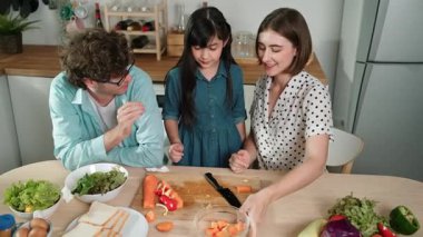 Çekici beyaz anne ve baba Asyalı kızıyla taze yemek pişirmek için birlikte vakit geçiriyorlar. Enerjik anne ve baba mutfakta salata yapmak için taze sebze hazırlıyor. Sağlıklı yiyecekler. Pedagoji.