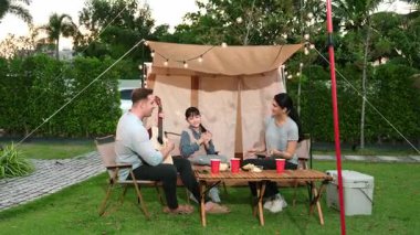 Baba gitar çalarken anne ve kız birlikte kampta oturuyorlar. Ebeveynler, genç nesil çapraz kuşak farklılıklarıyla zaman geçirmek için açık hava kamp aktivitelerini kullanıyorlar. Uyumsuzluk.