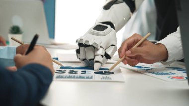 Bir insan ve robotik el, işbirlikçi bir iş toplantısına katılır, veri grafiklerini birlikte analiz eder, teknolojinin yenilikçi takım çalışmasındaki rolünü betimler. Noogenesis