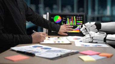 Bir insan eli, çağdaş bir ofiste robot bir elle takım çalışmasını ve yeniliğini teşvik etmek için renkli grafikler ve veri görselleştirmelerini analiz eder. Noogenesis