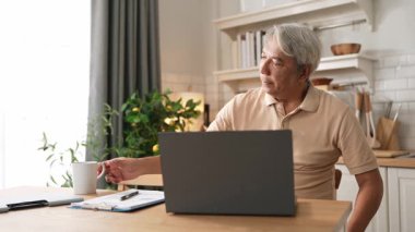 Zeki kıdemli iş adamı sıcak kahve içerken dizüstü bilgisayara bakıyor. Yaşlı proje yöneticisi finansal verileri kontrol ederken ve evde otururken karar veriyor. Uzaktan çalışma konsepti. Myrmidon.