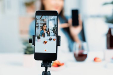 Seçici odaklı akıllı telefon ve gülümseyen genç, güzel Asyalı karışık çilek ve kiraz suyu detoksu değerlendirmesi sosyal medyada sağlıklı ürün tanıtımı sunuyor. Taktik.