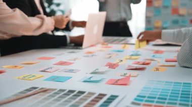 Parlak bir ofiste yapılan ve ellerin renkli notlar ve renk örnekleriyle fikirleri tartışarak takım çalışmasını ve yaratıcılığını sergilediği ortak bir toplantı. SACTR