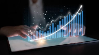 Bir iş adamı, modern bir ofis ortamında finansal büyüme ve pazar eğilimlerini gösteren holografik grafik grafikler ve grafikleri inceleyerek bir tabletle ilgilenir. Etki